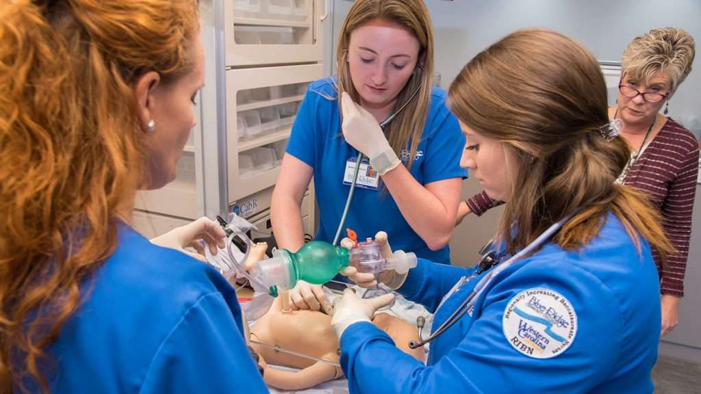 three nursing students work on a simulated infant. One of the students has a prominent R I B N patch on the shoulder of her uniform